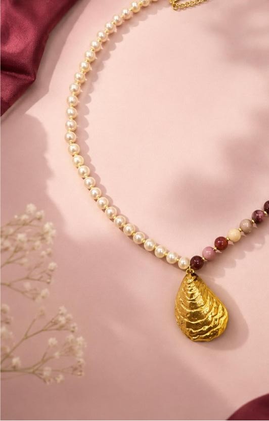 Aurore necklace — freshwater pearls graduating into burgundy and ochre stones with a hammered gold shell pendant, laid on blush paper with baby's breath