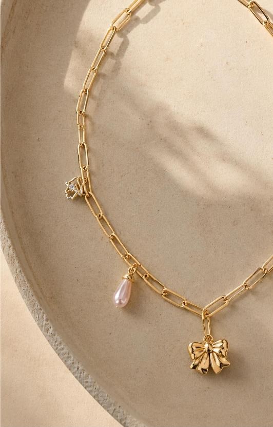 Constance — gold paperclip chain with ribbon bow, pink pearl drop and crystal star charms on sand