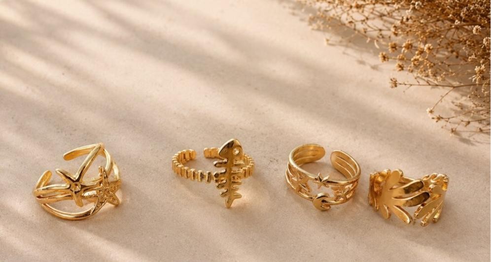 Marina — four gold stacking rings on sand: starfish, fish bone, star-and-chain, and palm, under dappled light